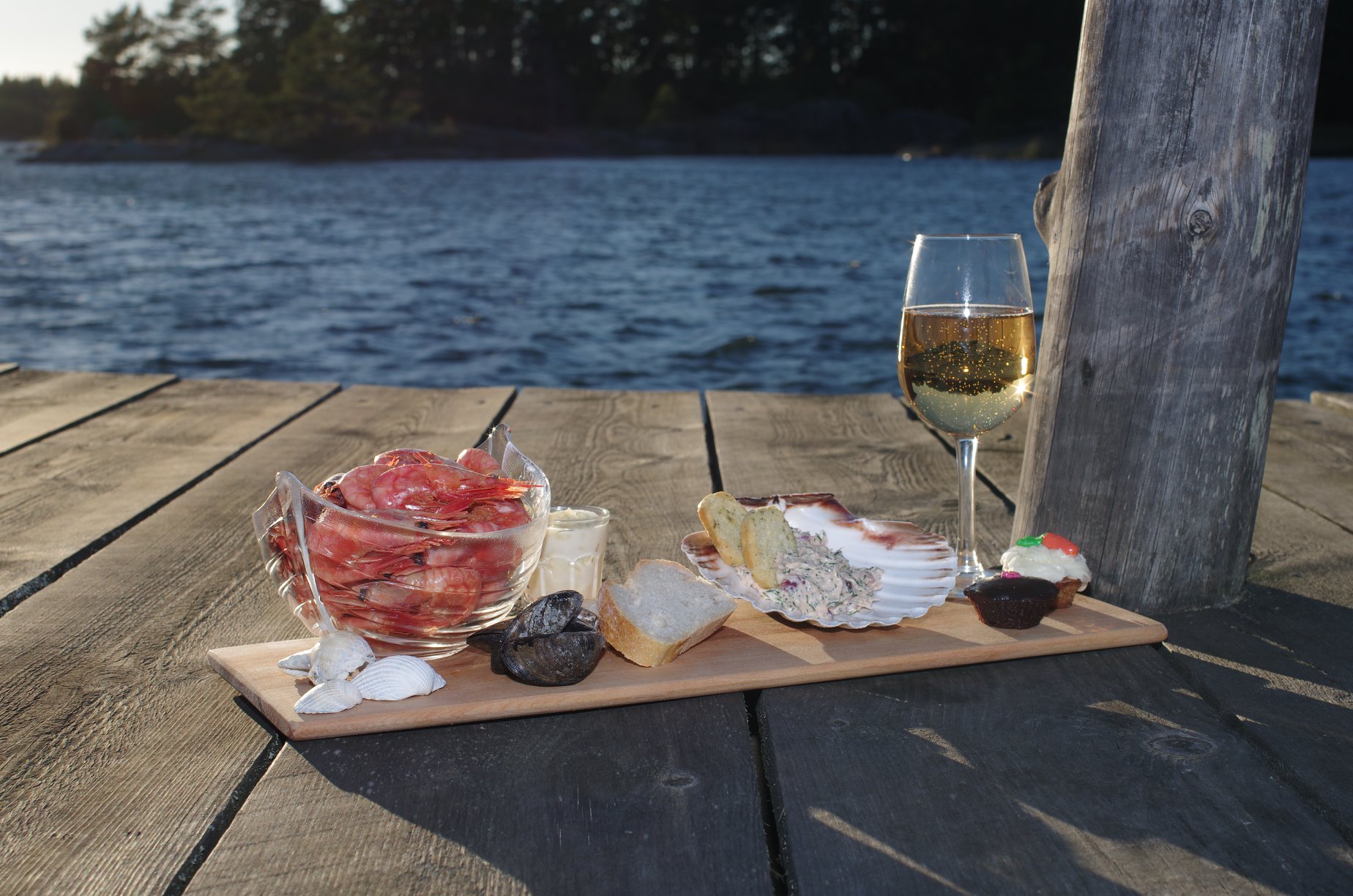 Räkor, ett glas vitt vin och bröd, tillsammans med snäckskal på en bricka vid vattnet