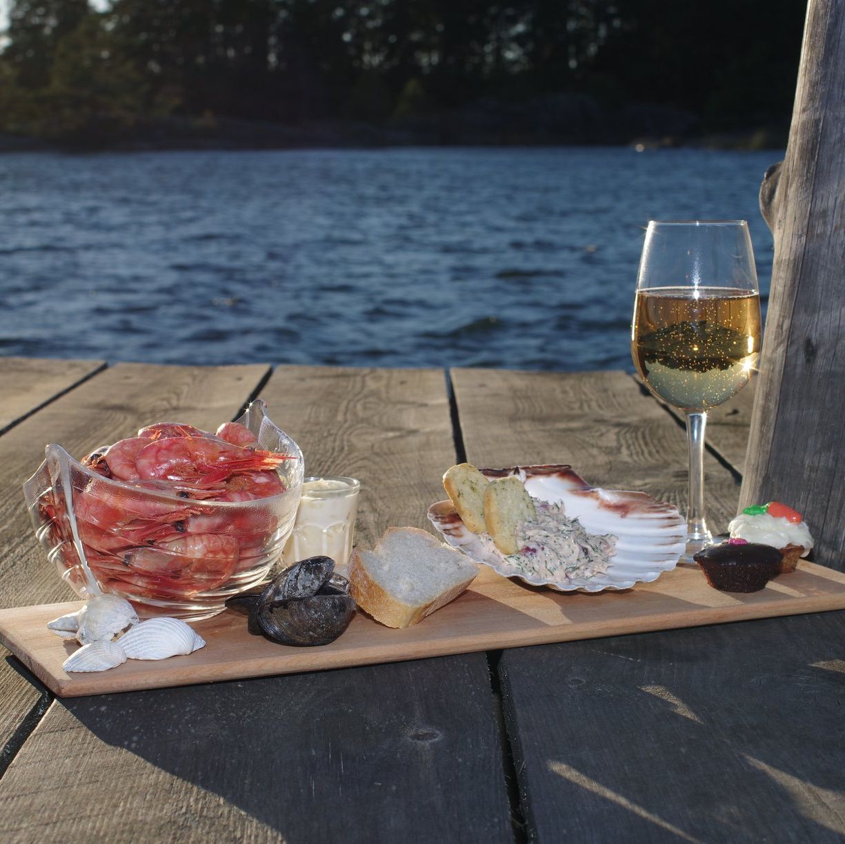 Räkor, ett glas vitt vin och bröd, tillsammans med snäckskal på en bricka vid vattnet